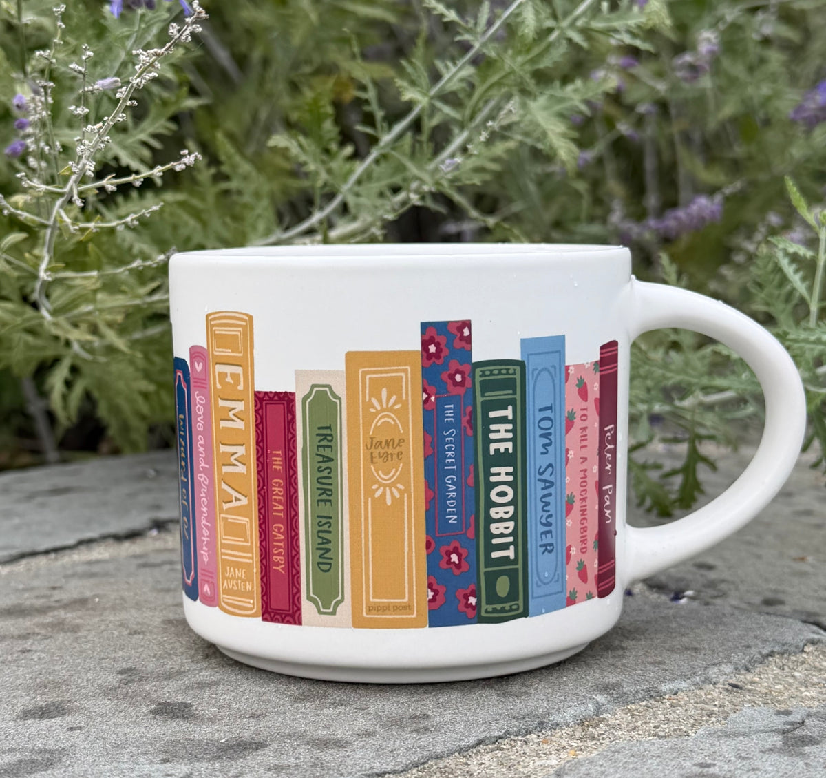 White mug with colorful classic book-spine illustrations on a stone surface in front of garden greenery.